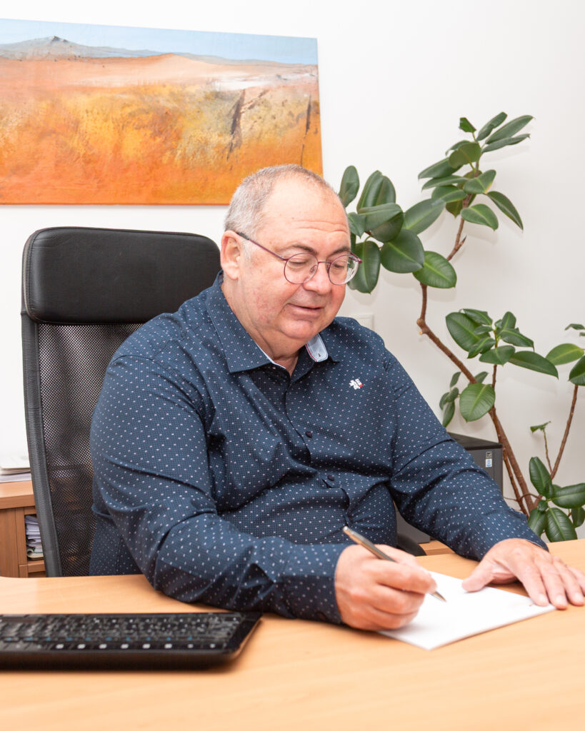 Mag. Walter Gusel (freundlicher Steuerberater) schreibt konzentriert auf Papier, lächelt leicht; im Hintergrund eine Pflanze und ein oranges Bild, professionelle Atmosphäre.