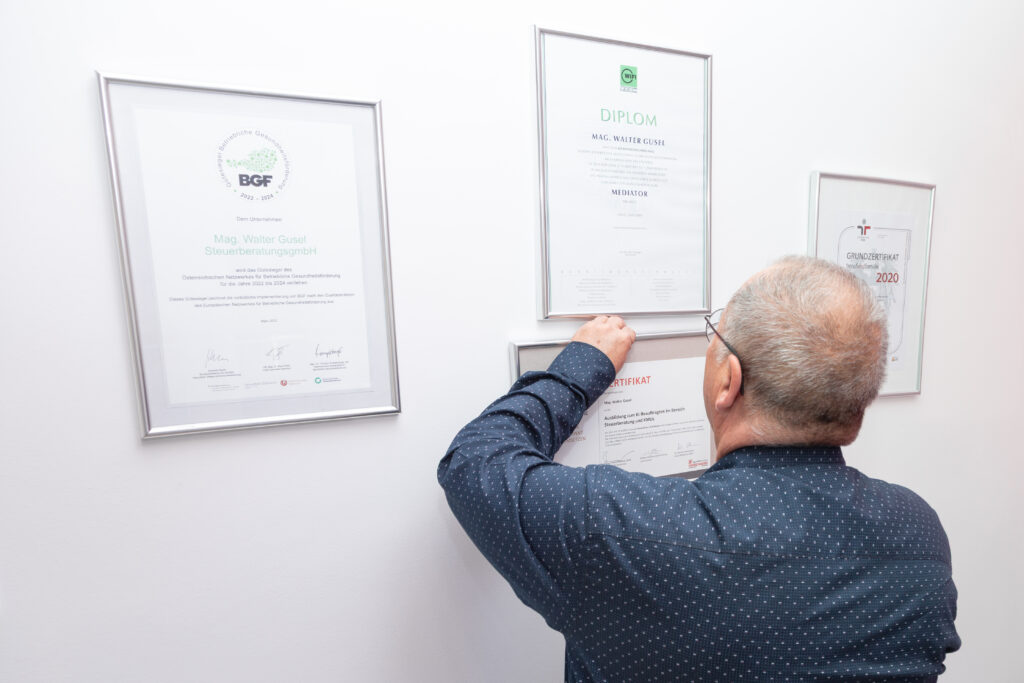 Steuerberater Mag. Walter Gusel bringt Zertifikate an einer Wand an und präsentiert stolz die erreichten Auszeichnungen. 