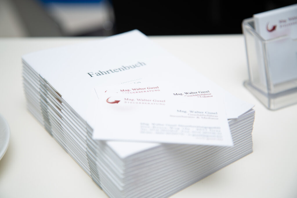 Mehrere Fahrtenbücher (gedruckt) liegen auf einem Bürotisch. Die Bücher sind geschlossen. In den Büchern werden tabellarische Einträge mit Datumsangaben, Fahrstrecken und Kilometerständen gezeigt.