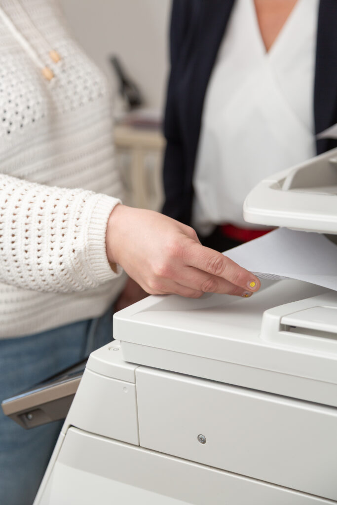 Zwei Hände arbeiten gemeinsam an einem Dokumentenscan: Die linke Hand (mit schlichtem Nagellack) hält eine Rechnung gerade am Scanner, während die rechte Hand den Deckel des Büroscanners bedient. Im Fokus liegt die professionelle Zusammenarbeit – die Blätter sind ordentlich ausgerichtet.