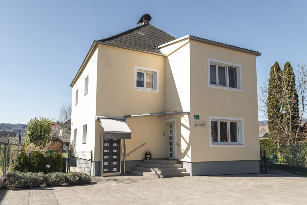 Ihre Anfahrt: Mehrstöckiges Bürogebäude mit markanter gelber Fassade und großen Fensterfronten. Der moderne Bau hat einen glatten Putz in Sonnengelb mit grauen Vor dem Eingang befindet sich ein gepflasterter Vorplatz. Über der gläsernen Eingangstür ist das Schild 'Steuerkanzlei Mag. Walter Gusel' in dunklen Buchstaben angebracht. Die Architektur wirkt professionell und einladend zugleich.